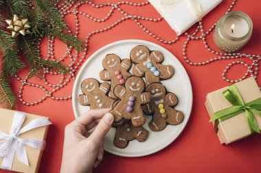 Beyaz bir tabakta zencefilli kurabiyelerle süslenmiş, etrafı tatil dekorlarıyla çevrili ve kırmızı arka planda hediyelerle dolu. Üst manzara, düz uzanma.