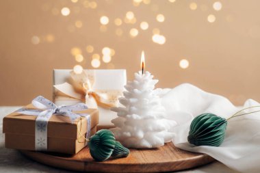 Beyaz yanan mum, hediye kutuları ve bej arka planda Noel ışıklarıyla süslü küçük kağıt bal peteği..