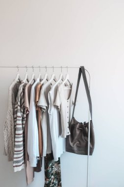 Minimalist giysi askılığı, düzgün düzenlenmiş nötr renkli giysiler ve açık arka planda şık bir deri çanta..