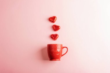 Kırmızı seramik kupa ve pastel pembe arka planda kalp şeklinde çikolatalar. Sevgililer Günü konsepti. Üst manzara, düz uzanma.