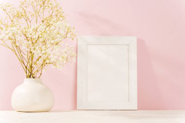 Beyaz vazo, bebek nefesi çiçekleri ve pembe bir duvara dayalı boş resim çerçevesi. Zarif güzelliği ve zarafeti simgeliyor. Bahar konsepti, model..