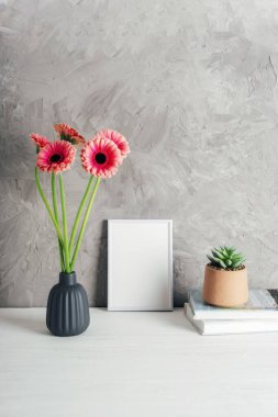 Beyaz resim çerçevesinin yanında gri seramik bir vazoda pembe gerbera çiçekleri ve modern iç plan üzerinde sulu bir saksı. Model.