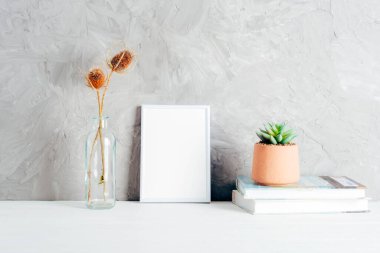 Boş beyaz çerçeveli minimalist ev dekoru, cam bir vazoda kurumuş çiçekler ve dokulu gri bir duvara karşı kitapların üzerinde sulu bir saksı. Model.
