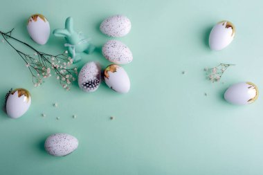 Beyaz ve altın renkli Paskalya yumurtaları, yeşil kadife bir tavşan ve pastel arkaplanda narin çiçekler. Üst görünüm, düz görünüm, kopyalama alanı.