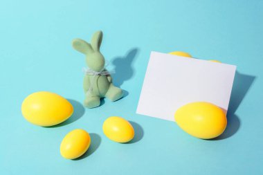 Yeşil kadife tavşanlı paskalya kompozisyonu, sarı yumurtalar, ve pastel mavi arka planda beyaz bir kart, bahar kutlaması ve bayram selamlarını simgeliyor. Model.