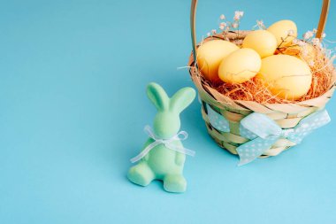 Sarı yumurtalı Paskalya sepeti ve pastel mavi arka planda kurdeleli yeşil tavşan heykelciği. Paskalya tebrik kartı. Boşluğu kopyala.