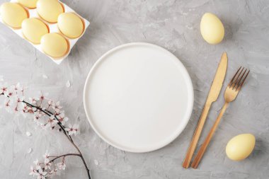 Altın çatal bıçaklı boş beyaz tabak, sarı yumurta ve gri arka planda kiraz çiçeği. Paskalya konsepti. Üst görünüm, düz görünüm, model.
