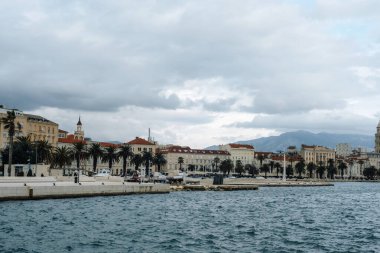 Bulutlu gökyüzünün altında tarihi binalar ve palmiye ağaçları olan deniz kenarı geçidi. Ayrıl, Hırvatistan.