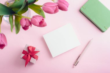 Vazoda pembe laleler, boş kart, not defteri ve pastel pembe arka planda hediye kutusu. Bahar tatili konsepti. Üst görünüm, model.