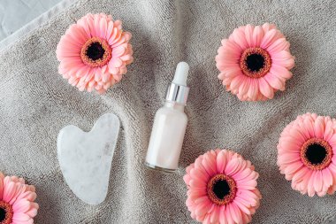 Serum şişesi ve yumuşak havlunun üzerinde pembe gerbera olan gua sha taşı. Cilt bakımı. Üst manzara, düz uzanma.