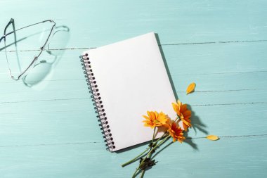 Turuncu çiçekli, turkuaz ahşap arka planda gözlüklü boş spiral defter. Üst görünüm, düz görünüm, model.