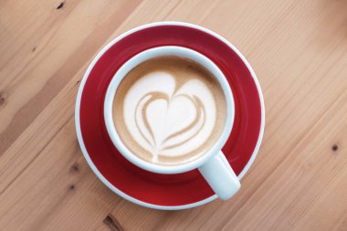 Kırmızı çay tabağı ve ahşap masa üzerinde kalp şeklinde latte sanatı olan kahve fincanının en üst görüntüsü. Üst manzara, düz uzanma.