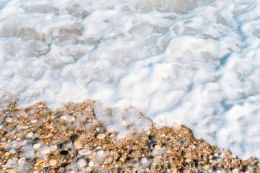 Sakin bir sahil şeridinde yumuşak çakıl taşlarının üzerinden yavaşça geçen deniz köpüğü doğal dokuyu ve kıyı sükunetini yakalıyor. Doğal arkaplan, üst görünüm.
