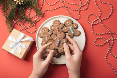 Elleri beyaz bir tabakta zencefilli kurabiye tutuyordu. Renkli süslemelerle süslenmiş ve kırmızı arka planda hediyeler vardı. Üst görünüm.