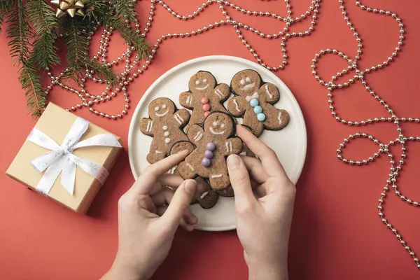 Elleri beyaz bir tabakta zencefilli kurabiye tutuyordu. Renkli süslemelerle süslenmiş ve kırmızı arka planda hediyeler vardı. Üst görünüm.
