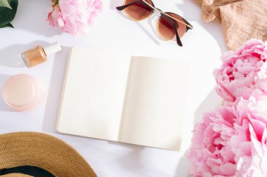 Beyaz arka planda pembe şakayıklar, güneş gözlükleri, oje ve hasır şapkayla çevrili boş sayfaları olan açık bir defter. Üst görünüm, düz görünüm, model.