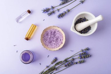 Bir kasede lavanta banyosu tuzu, lavanta çiçekli harç ve kabuk, mor arka planda aroma yağı ve hidrolat. Doğal bitkiler ilaç, aromaterapi konsepti. Üst manzara, düz uzanma.