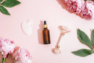 Kozmetik serum şişesi, yüz silindiri, gua sha aleti ve pembe arka planda pembe şakayıklar, cilt bakımı. Üst manzara, düz uzanma.
