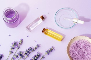 Kâsede lavanta banyosu tuzu, lavanta çiçekleri, aromatik yağ, petri kabında yüz serumu pipetli ve mor arka planda hidrolat. Doğal bitkisel ilaçlar, aromaterapi konsepti. Üst manzara, düz uzanma.