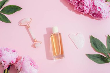 Kozmetik şişe, yüz silindiri, gua sha taşı ve pembe arka planda pembe şakayıklar, cilt bakımı konsepti. Üst manzara, düz uzanma.