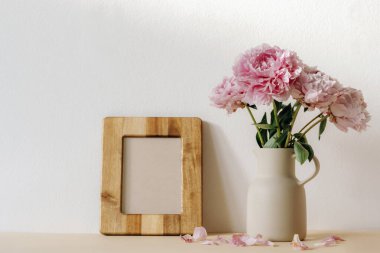 Seramik sürahide pembe şakayıklar ve beyaz duvara dayalı boş ahşap çerçeve. Model.