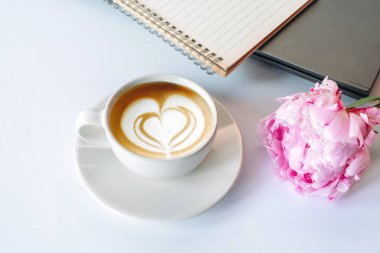 Beyaz arka planda bir fincan kapuçino, defter ve şakayık çiçeği. Üst görünüm.