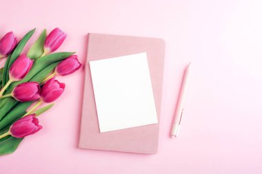 Beyaz kart, pembe laleler, not defteri ve pastel pembe arka planda kalem. Üst görünüm, düz görünüm, model.