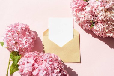 Kraft zarfında, pastel arka planda pembe ortanca çiçekleri olan boş beyaz kart. Üst görünüm, düz görünüm, model.