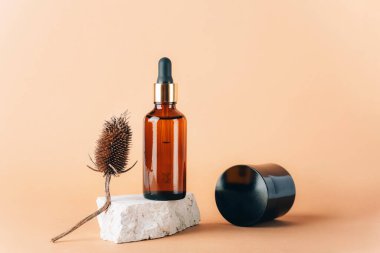 Doğal taş podyumda kahverengi kozmetik serumu şişesi, devedikeni dolgunluğu ve doğal bej arka planda krema kavanozu. Yakın çekim..