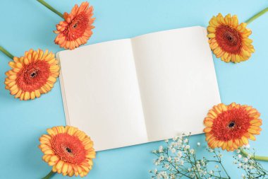 Mavi arka planda boş defter ve turuncu gerbera çiçekleri. Yaz tatili, tebrik kartı. Üst görünüm, model.