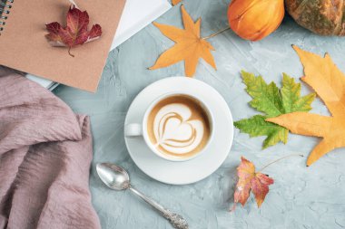 White cup with latte art heart surrounded by autumn leaves, pumpkins and notebook on textured background. Autumn concept. Top view, flat lay.