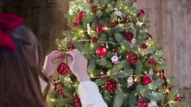 Saçında kırmızı bir fiyonk olan, Noel ağacına kırmızı oyuncaklar asan küçük bir kız resmi. Büyük ağaç, oyuncaklar ve beyaz ışıklarla dolu. Kış tatili kavramı