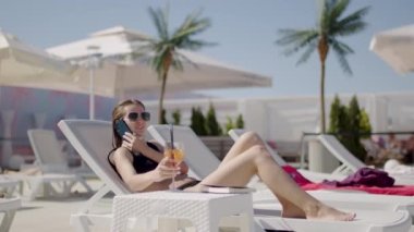 Genç ve güzel bir kadın telefonda konuşuyor. Güneşli bir yerde uzanıp kokteyl içiyor.