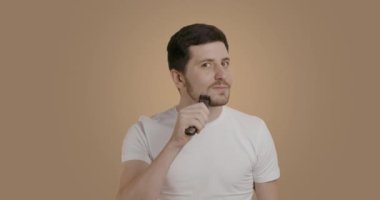 Koyu saçlı sakallı adam çenesindeki kesik saçı tıraş etmek için elektrikli makas kullanıyor. Genç adam tek başına sakalıyla ilgileniyor. Evde kolay tıraş olma kavramı