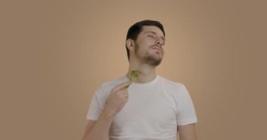 Temiz ve taze derisi olan genç ve yakışıklı bir adam yeşim taşından bir masaj aleti kullanarak boynuna masaj yapıyor. Genç bir adamın sağlıklı yaşam tarzı. Kırışıklık önleyici tedavi ve cilt bakımı kavramı.