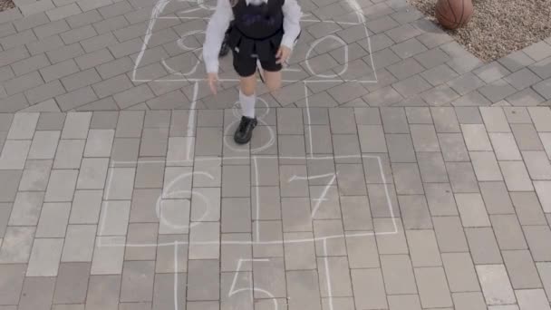 地面に白いチョークで描かれたインタラクティブなゲーム 制服を着た女子学生が地面に描かれた数字を飛び越える 学期の概念 — ストック動画