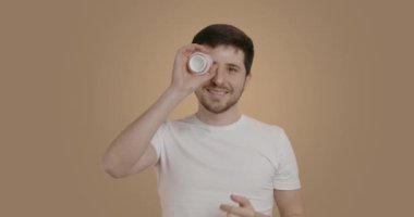 Nice man in a white t-shirt doing his hair care procedures. Brunette man who applied a moisturizing cream and then put the container to his eyes, smiling widely at the camera. Care concept
