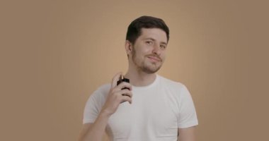 A man dressed in a white T-shirt on a beige studio background puts on perfume and smiles widely at the camera. A young man who perfumes himself several times in the neck area.