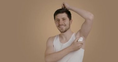 Man with a big smile who looks after himself in the bathroom. Young man applying ball deodorant under his arm. Brunette man with a beard doing his morning, evening grooming routine.