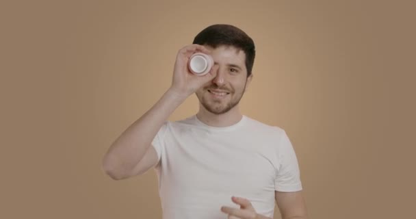 白いTシャツの素敵な男がヘアケア手順を実行しています 保湿クリームを塗り 容器を目につけ カメラで大きく笑顔を浮かべるブルネットの男 介護の概念 — ストック動画
