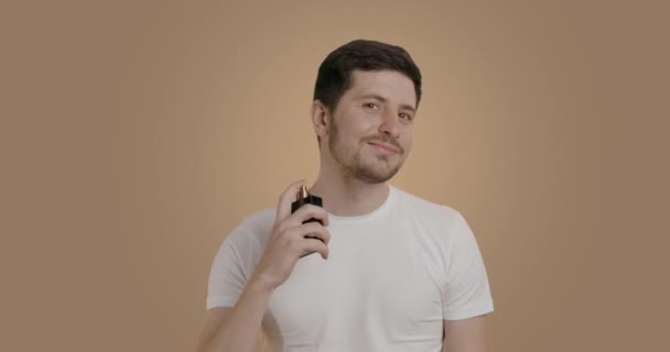 一个身穿白色T恤 背景是米色工作室的男人在镜头前放上香水 面带微笑 一个年轻人 在颈部区域给自己喷了好几次香水 — 图库视频影像