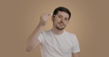 Smiling man doing his morning,evening grooming procedures. Young man applying a moisturizing serum to the eye area with a pipette. Brunette man with a beard cleaning himself in the bathroom.
