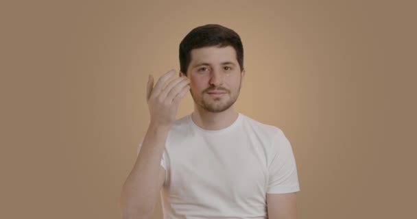 布鲁内特男子在工作室背景 同时小心地按摩他的眼部区域 他的手指 一个留着胡子 穿着白色T恤的年轻人 用润肤霜护理他的脸 — 图库视频影像