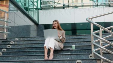 Stylish student sitting on the stairs in front of a modern university, giving an online speech and explaining with her hands. Student during online classes outside in the open air. Student life