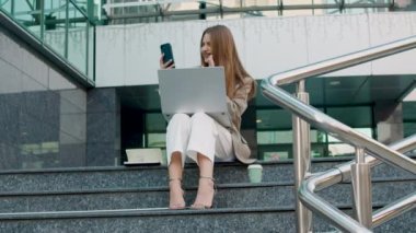 Stylish and cheerful student talks on the phone with her parents and shows them hello. Student at break time sitting on the stairs outside in front of the university working on the computer.