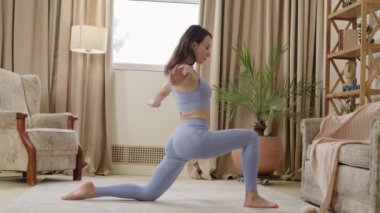 Side view of young woman with athletic body wearing blue sportswear doing leg stretching yoga exercise on fitness mat in bright living room. Concept of health care and mental therapy at home