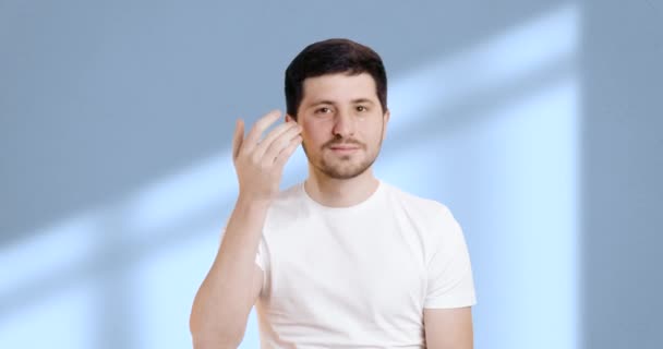布鲁内特男子在浴室的蓝色背景上 同时小心地用手指按摩他的眼部区域 穿着白色T恤 用润肤霜做面部护理的年轻人 — 图库视频影像