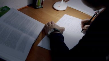 Öğrenci tezini bir lamba ışığında yazmaya hazırlanıyor. Defterine birkaç fikir yazan ve onun materyallerini çizen bir çocuğun yakın plan resmi. Öğrenci konsepti