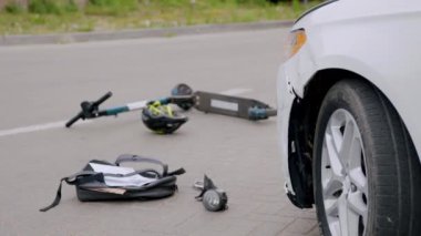 Lüks beyaz bir araba ile asfalt yolda elektrikli scooter arasında bir trafik kazası. Bisikletçilerin kaskları, sırt çantaları ve araba parçaları yere atıldı. Kaza kavramı.