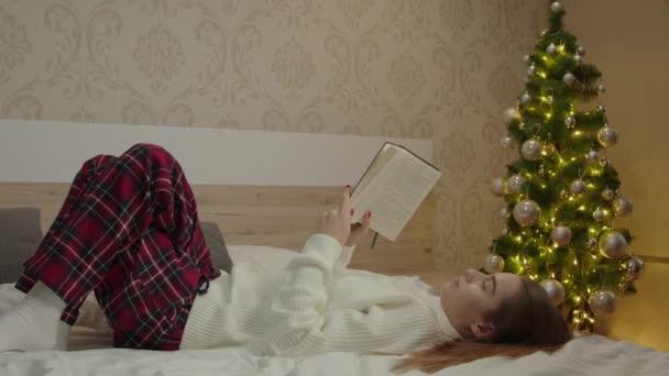 就寝時間 休息のコンセプト 夜のクリスマスのコンセプトでベッドに座って読書本を読んでいるパジャマの女性 — ストック動画